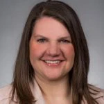 A woman with straight brown hair, wearing a light-colored blazer, smiles at the camera against a plain gray background.