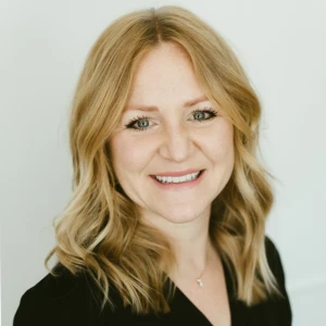 A woman with shoulder-length blonde hair and a black top smiles at the camera against a plain light background.