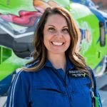 Woman in a blue jumpsuit smiling in front of a green helicopter with colorful designs.