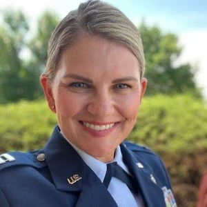 A woman in a U.S. military uniform smiles outdoors, with greenery and a blue sky in the background.