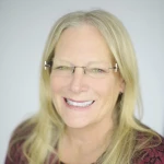A woman with long blonde hair and glasses is smiling at the camera, wearing a patterned top against a plain light background.