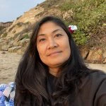 A woman with long dark hair sits on a beach near rocky cliffs, wearing a dark top and a flower in her hair, looking at the camera.