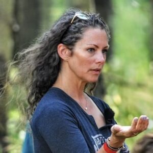 A woman with long curly hair and sunglasses on her head stands outdoors, gesturing as if addressing BCEN conference speakers, while looking to the side against a backdrop of blurred greenery.