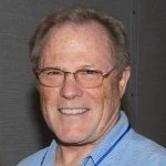 A middle-aged man with short light hair and glasses smiles at the camera. He is wearing a light blue collared shirt and standing in front of a neutral background.