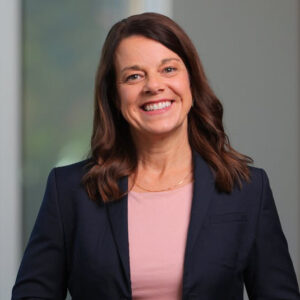 A woman with long brown hair wearing a navy blazer and light pink top smiles while standing indoors near a window, ready to join fellow BCEN conference speakers.