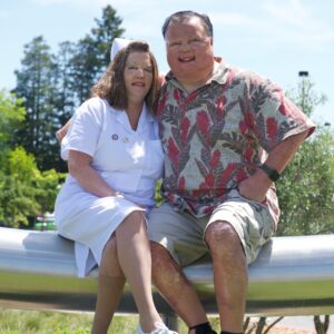 A woman in a white nurse uniform and a man in a floral shirt and shorts, BCEN conference speakers, sit together on a metal beam outdoors, smiling at the camera with trees and greenery in the background.