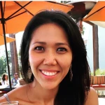 A woman with long dark hair smiles at the camera while sitting outdoors under orange umbrellas.