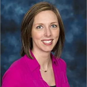 Woman with straight, light brown hair wearing a bright pink blouse, smiling at the camera with a blue, patterned background.