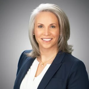 A woman with shoulder-length gray hair, wearing a navy blazer and white blouse, smiles at the camera against a plain gray background.