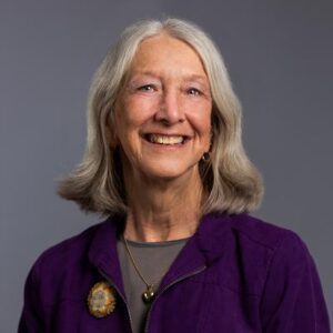 Older woman with gray hair, wearing a purple jacket and a gray top, smiling in front of a plain gray background—one of the distinguished BCEN conference speakers.