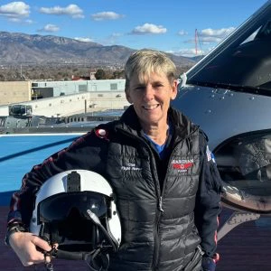 A person in a flight suit holds a helmet while standing next to a helicopter on a rooftop helipad with mountains in the background.