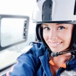 A person wearing a helmet and uniform smiles while sitting inside a helicopter cockpit.