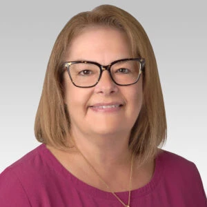 A woman with straight, shoulder-length light brown hair, glasses, and a maroon top smiles at the camera against a plain light gray background.