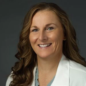 Woman with long, wavy brown hair wearing a white lab coat smiles at the camera against a plain dark background.