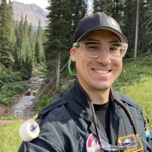 A person in outdoor gear and clear protective glasses smiles for a selfie in a forested area with a stream and mountains in the background, reminiscent of BCEN conference speakers enjoying nature between sessions.