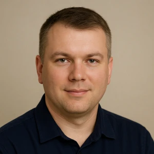 A man with short brown hair and light skin wears a dark blue collared shirt and faces the camera, smiling slightly against a plain beige background.