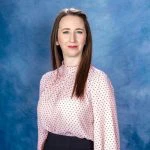 Woman with straight brown hair wearing a pink blouse with red polka dots and dark pants poses in front of a blue studio background.