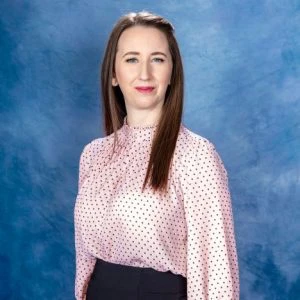 A woman with straight brown hair, wearing a light pink blouse with small black dots and dark pants, stands in front of a blue textured background.