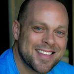 A smiling man with a shaved head and trimmed beard wearing a blue shirt, photographed in close-up.