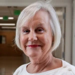 An older woman with short white hair and a white top stands indoors, smiling gently at the camera. The background appears to be a hallway.