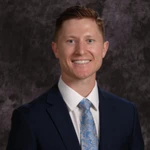 A man in a dark suit, white shirt, and patterned light blue tie smiles in front of a dark, textured background.