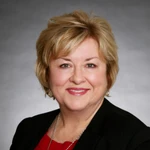 Portrait of a middle-aged woman with short blonde hair, wearing a black blazer over a red top, smiling at the camera against a gray background.