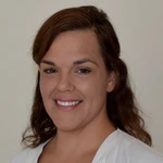 A woman with wavy brown hair, wearing a white top, is smiling at the camera against a plain light background.