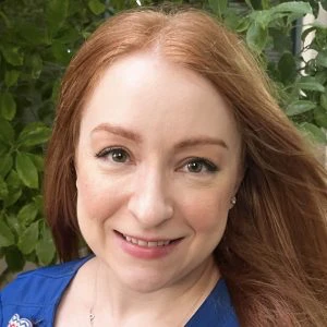 A woman with long red hair smiles at the camera, standing outdoors in front of green leafy plants, wearing a blue top.