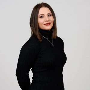 Woman with straight brown hair wearing a black turtleneck sweater and layered necklaces, standing against a plain light background.