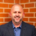 A man in a suit jacket and collared shirt stands smiling in front of a brick wall.