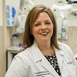 A woman in a white lab coat stands in a medical setting, smiling at the camera. Medical equipment and supplies are visible in the background.