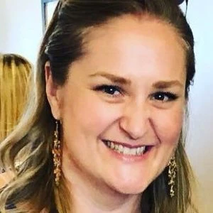 A woman with light brown hair and long earrings smiles at the camera in an indoor setting.