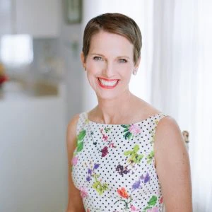 A woman with short brown hair, wearing a white sleeveless dress with a floral and polka dot pattern, smiles while seated indoors near a window.