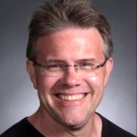 A man with short brown hair and glasses is smiling at the camera against a dark grey background.