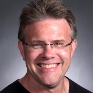 A man with short, grayish hair and glasses smiles at the camera. He is wearing a black shirt and the background is plain and dark.