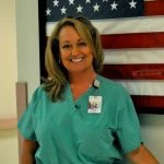 A woman in medical scrubs stands in front of a U.S. flag, smiling at the camera. She is wearing an ID badge.