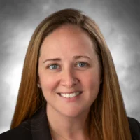 A woman with long light brown hair, wearing a dark blazer, is smiling in front of a gray, softly blurred background. She is the 2026 Distinguished TCRN Award Winner.