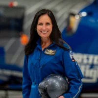 A woman in a blue flight suit stands indoors, holding a helmet and smiling at the camera. A helicopter is blurred in the background, highlighting her achievement as the 2026 Distinguished CFRN Award Winner.