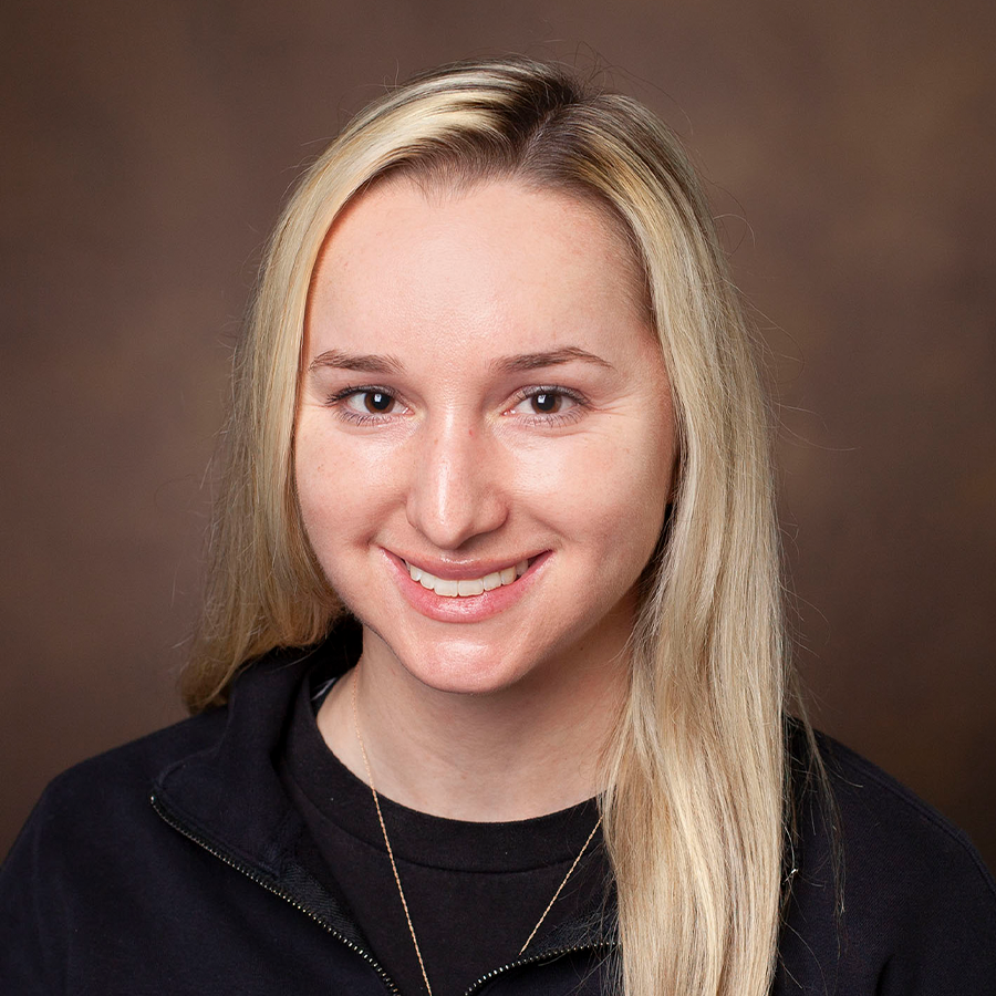 A woman with straight, long blonde hair wearing a black top and a necklace smiles in front of a plain brown background.
