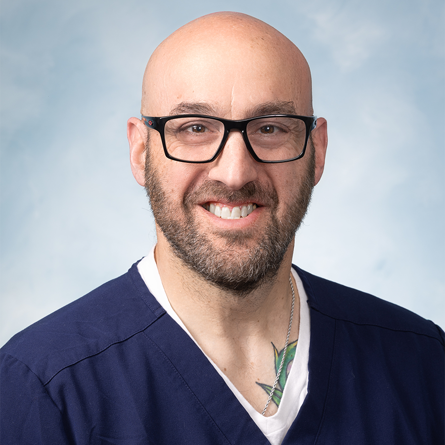 A bald man with glasses and a beard, wearing navy blue scrubs and a necklace, smiles at the camera against a light blue background.