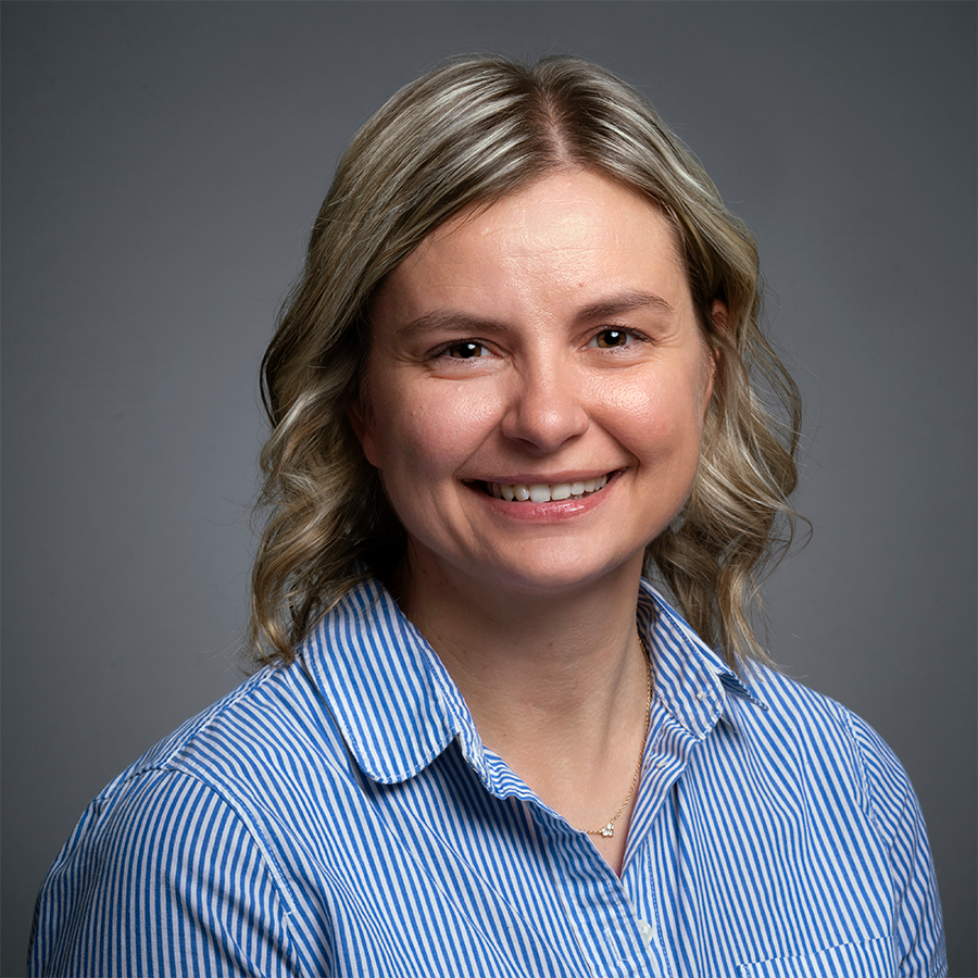 A woman with shoulder-length blond hair smiles at the camera. She is wearing a blue and white striped shirt and is posed against a plain gray background.