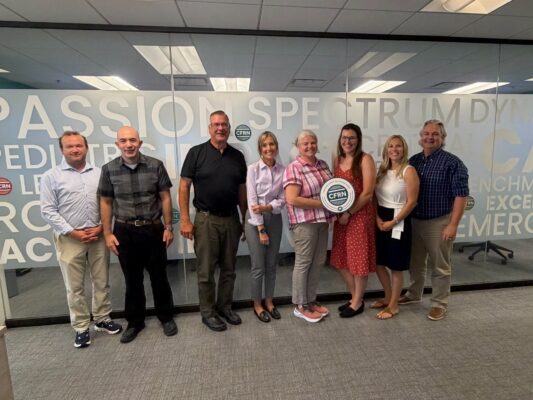 Eight people stand in a row indoors, some holding a round CFN sign, in front of a glass wall with large text in a modern office setting, gathering to discuss the latest CFRN exam updates.