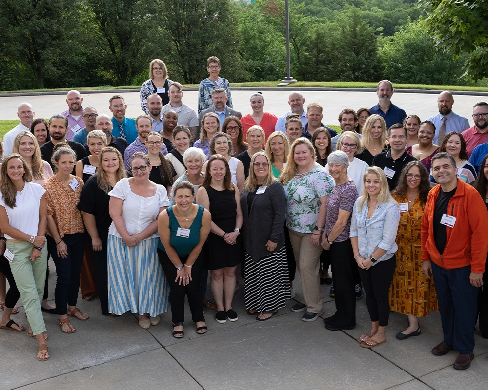 A large group of adults pose together outdoors on a paved area, with trees and greenery visible in the background.