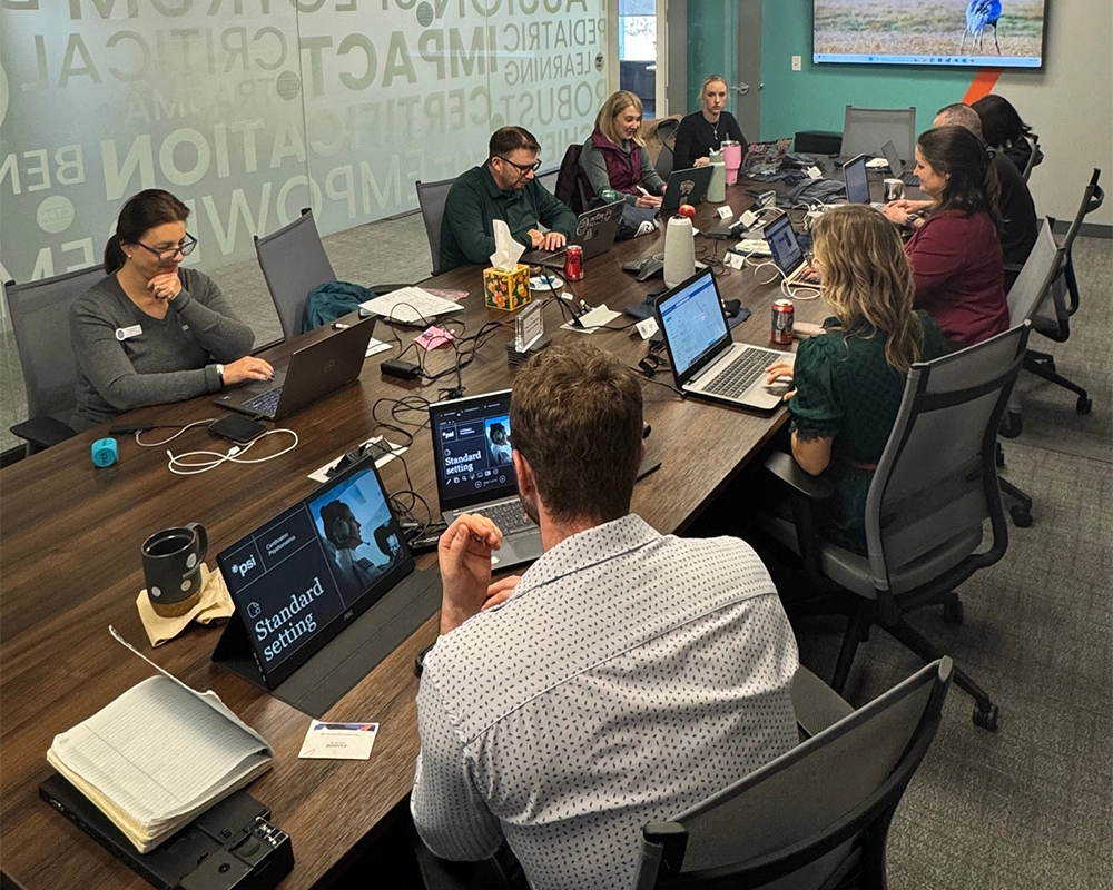 Seven people are seated around a conference table, working on laptops and paperwork in a modern office meeting room.