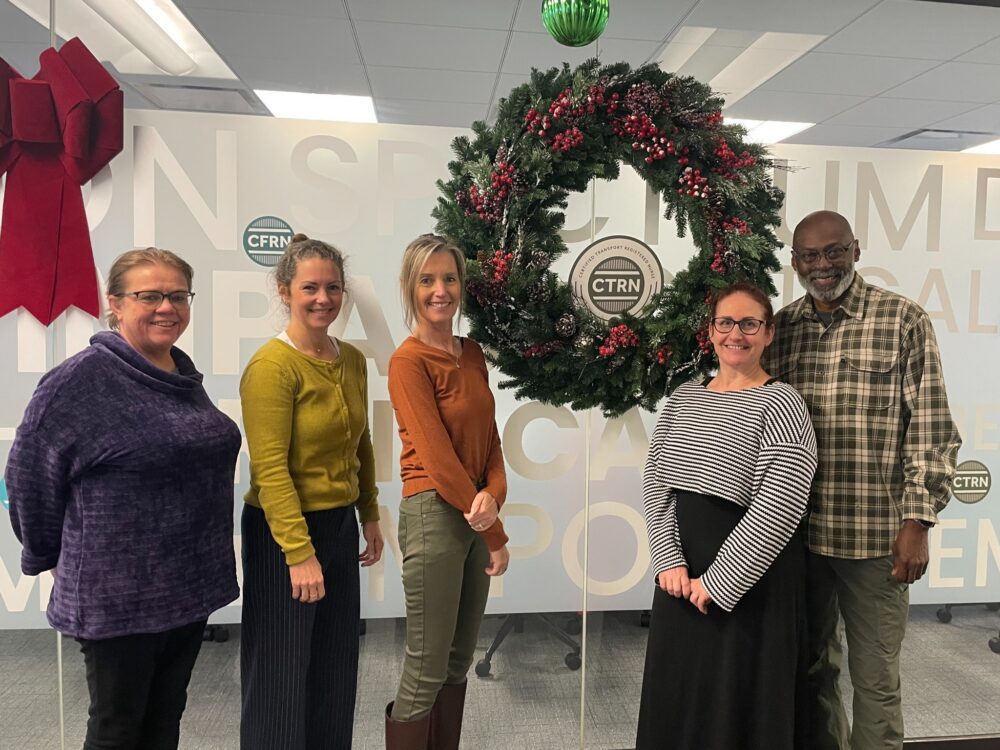 Five people stand in front of a large holiday wreath and office wall with "CTRN" logos and lettering, posing for a group photo.