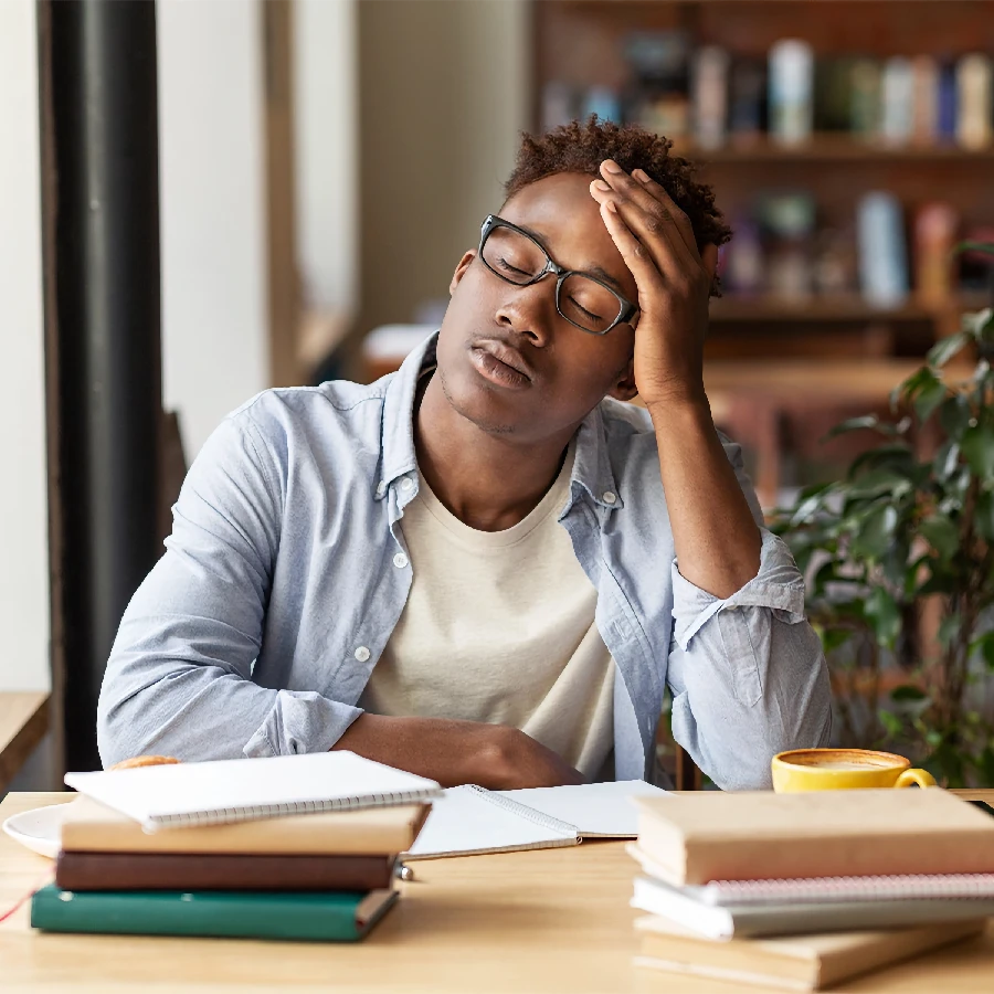 A person sitting at a desk with notebooks and a coffee cup, resting their head on their hand with eyes closed, appearing tired or stressed.