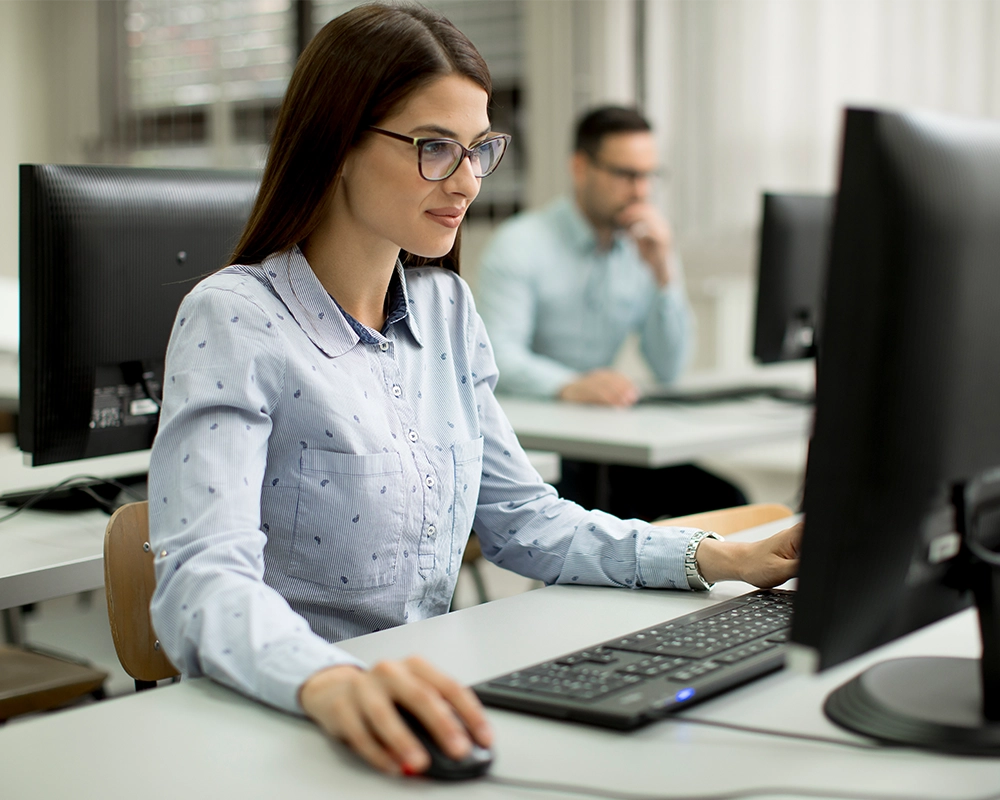 A woman using a desktop computer in an office setting, with another person working at a computer in the background.