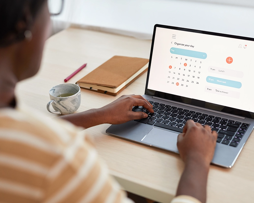 Person using a laptop to organize their day on a digital calendar, with a notebook and a cup nearby on a wooden desk.