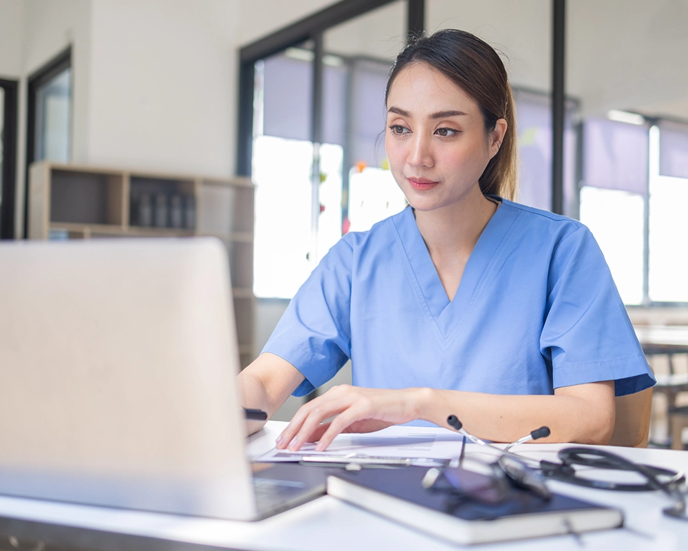 A woman in blue medical scrubs sits at a desk working on a laptop with papers, a pen, and a stethoscope nearby.