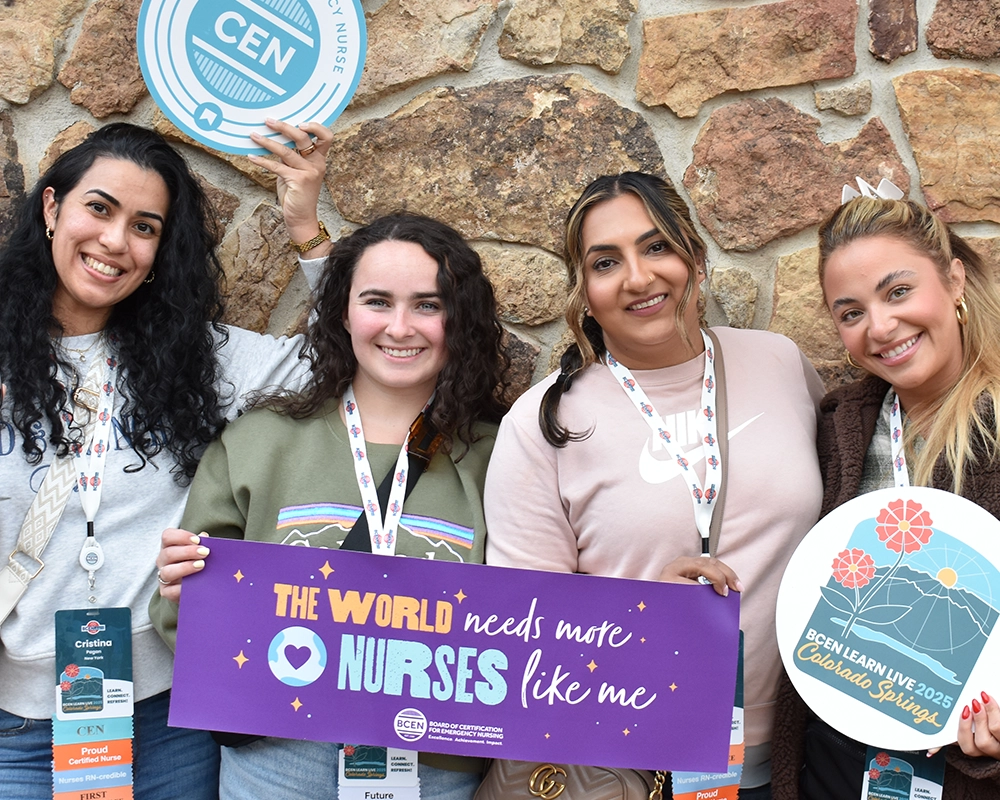 Four women smile at the camera, holding signs—including one that reads "The world needs more nurses like me"—while attending an emergency nursing conference in front of a stone wall.
